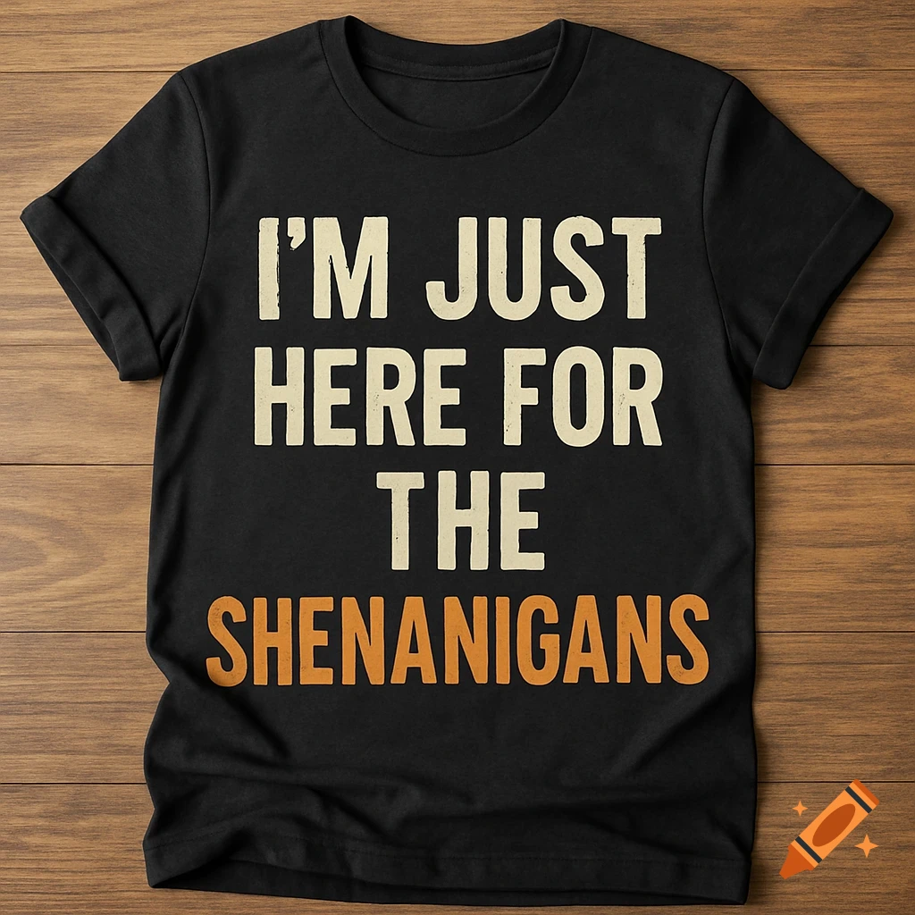 A black t-shirt with the quote 'I'm Just Here for the Shenanigans' in cream and orange lettering, laid flat on a wooden surface.