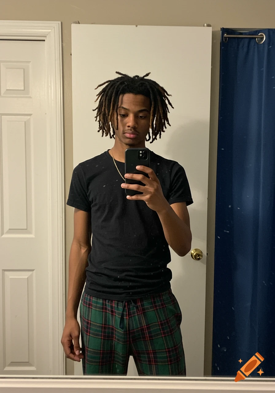 A young man with dreadlocks takes a mirror selfie in a black t-shirt and plaid pajama pants.