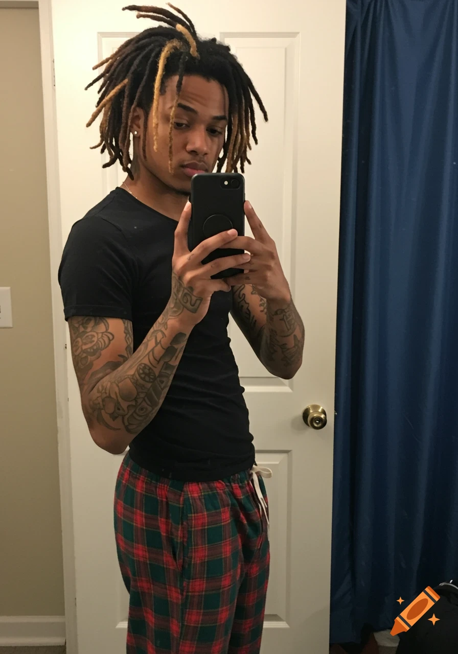 A young man with dreadlocks and arm tattoos takes a mirror selfie, wearing a black t-shirt and red and green plaid pajama pants.