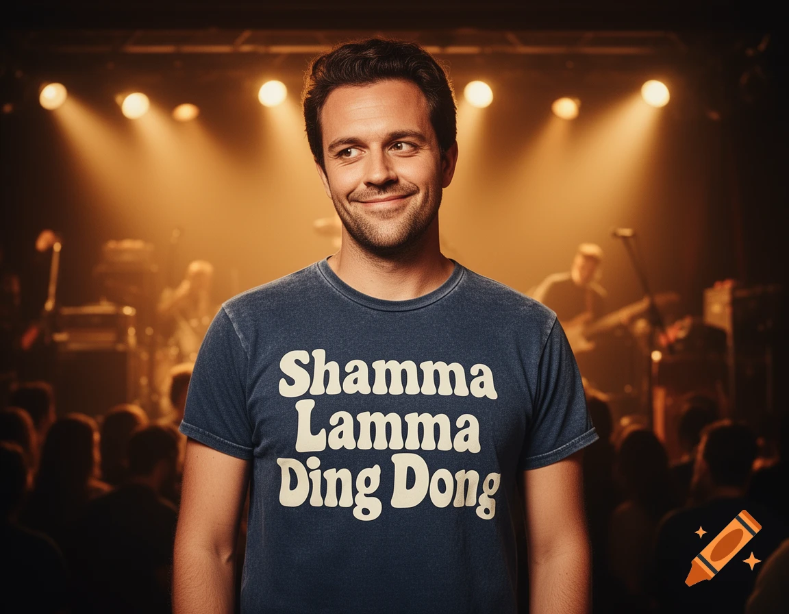 Smiling man in a blue 'Shamma Lamma Ding Dong' t-shirt on a stage with warm spotlights and blurred musicians.